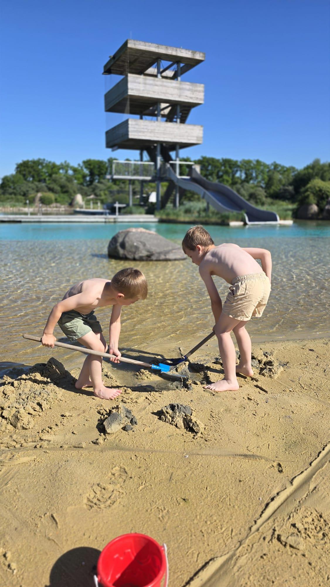 Zwei Kinder buddeln im Sand des Nicht-Schwimmer-Bereichs im Naturbad Olfen. Im Hintergrund ist der Sprung- und Aussichtsturm des Bades zu sehen.