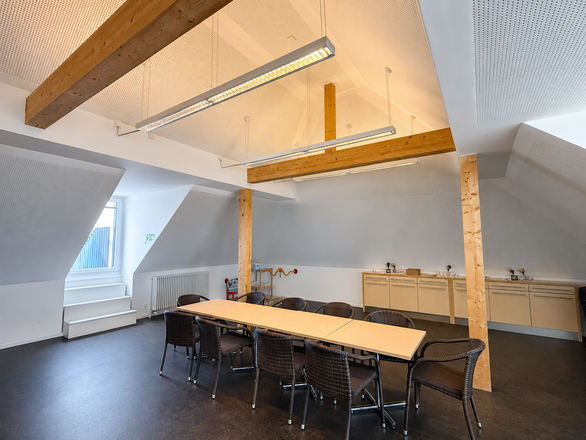 Heller Raum im Dachgeschoss mit sichtbaren Holzbalken, langen Hängeleuchten, einem großen Tisch mit Stühlen sowie Schränken an der Wand.