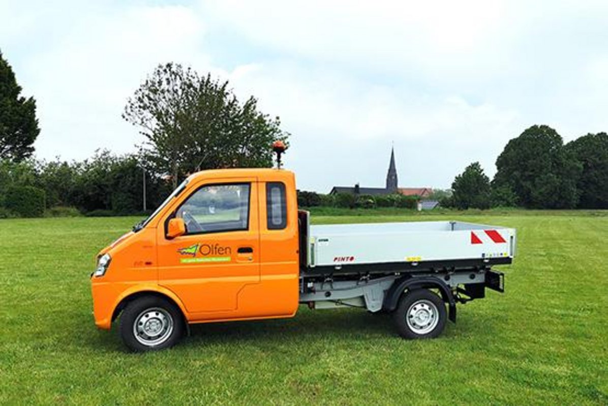 Ein orangener Pritschenwagen der Stadt Olfen steht auf einer grünen großen Fläche.