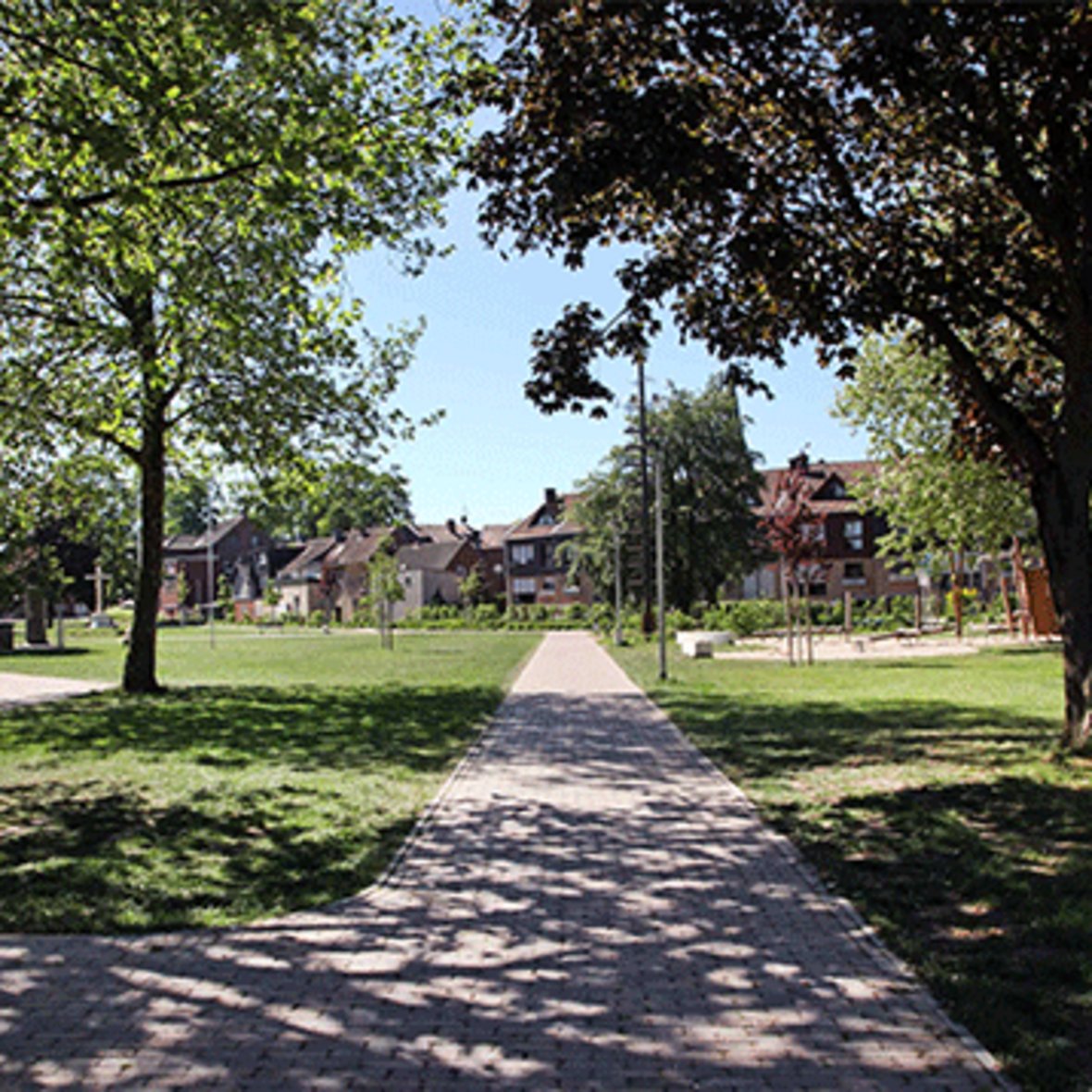 Zu sehen ist der gepflasterte Weg, der in den Olfener Stadtpark führt. Drumherum viele grüne Bäume und grüner Rasen. Außerdem ist im Hintergrund der Spielplatz zu erkennen.