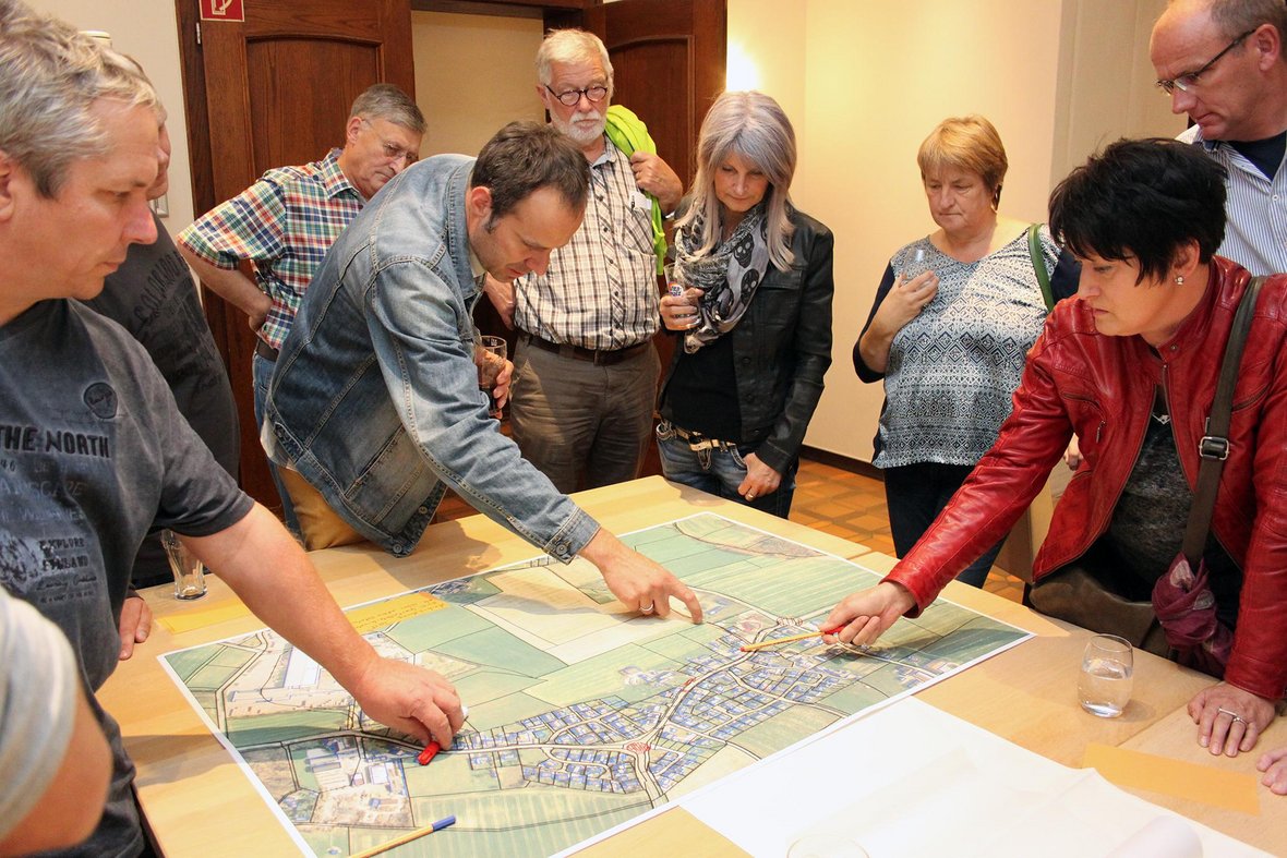 Auf dem Foto sind 8 Personen zu sehen, die um einen Tisch mit vielen Plänen herum stehen und Ideen und Notizen aufschreiben. Hier werden über Verbesserungsmöglichkeiten für den Ortsteil Vinnum gesprochen.