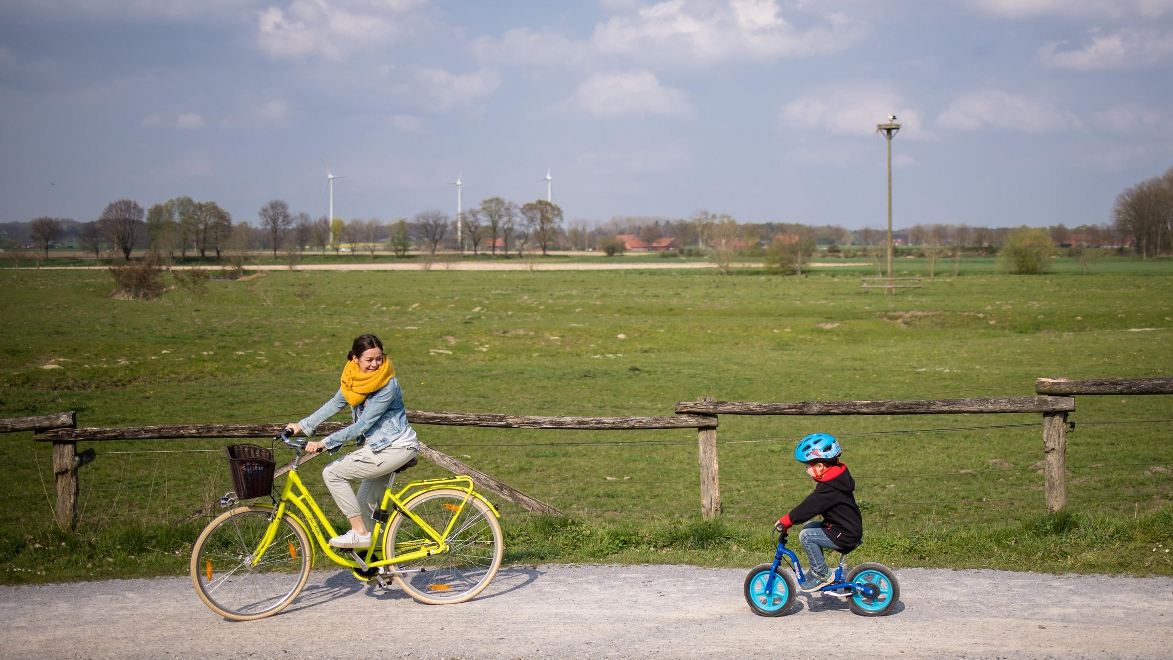 Eine Frau auf einem gelben Fahrrad fährt einen Weg durch die Steveraue Olfen. Hinter ihr fährt ein kleiner Junge mit seinem Laufrad. Im Hintergrund ist der Storchenhorst zu sehen.