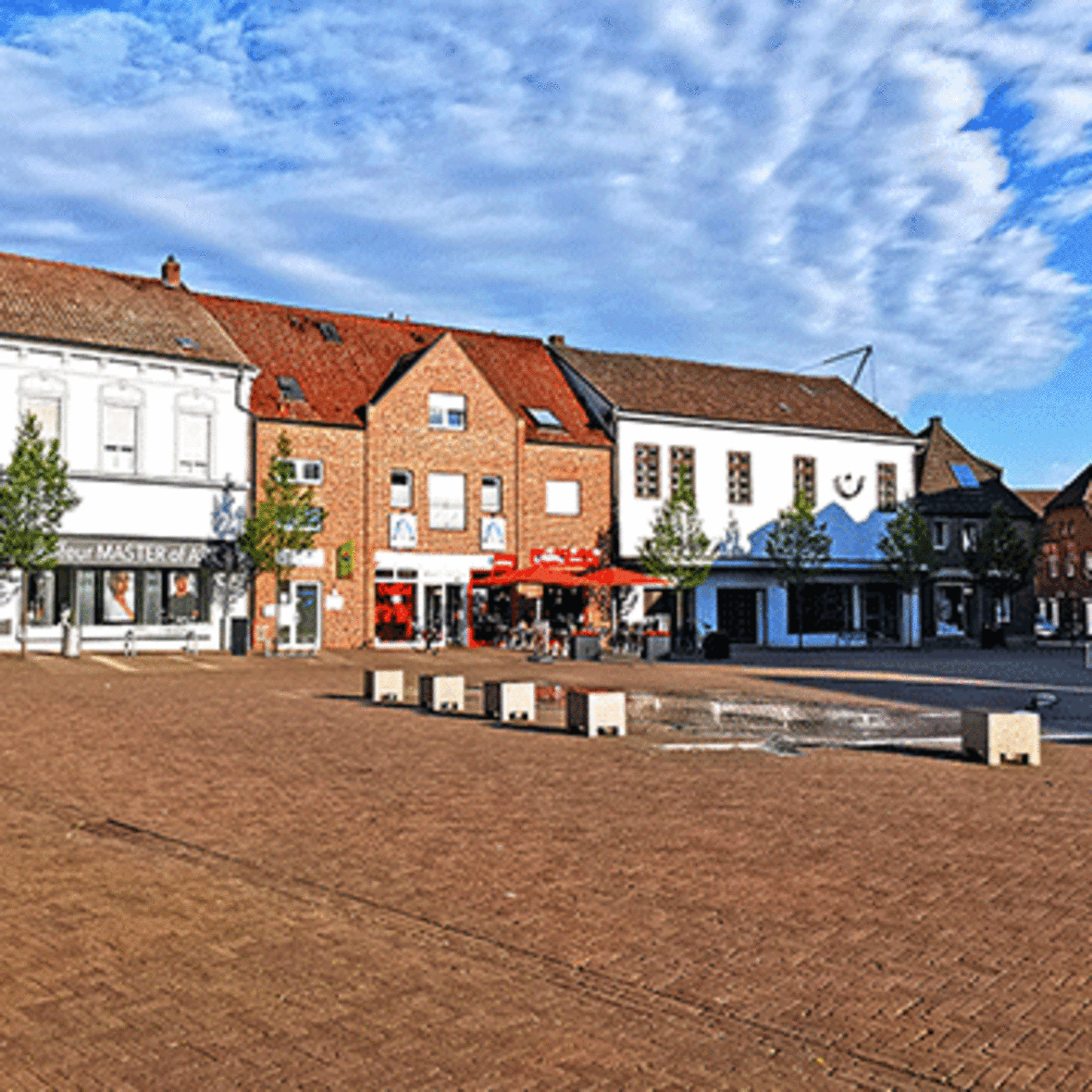 Als Symbol für die Unterseite des Einzehalndeskonzepts der Stadt Olfen ist der Marktplatz der Stadt Olfen zu sehen. Im Vordergrund der Brunnen, im Hintergrund sieht man mehrere Gebäude, u. a. der Eingang zur Marktpassage. 