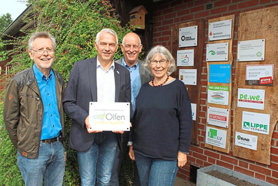 Bürgermeister Wilhelm Sendermann und drei Personen vom Biologischen Zentrum stehen an der Mitgliedertafel des Biologischen Zentrums. Herr Sendermann hält das Schild mit dem Logo der Stadt Olfen in der Hand.
