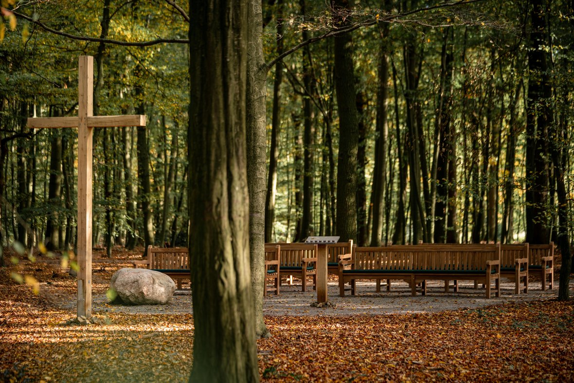 Auf dem Andachtsplatz steht ein großes Kreuz aus Holz und einige Bänke aus Holz zum Platznehmen. Drumherum sind viele Bäume zu erkennen. Der Platz befindet sich mitten im Wald.