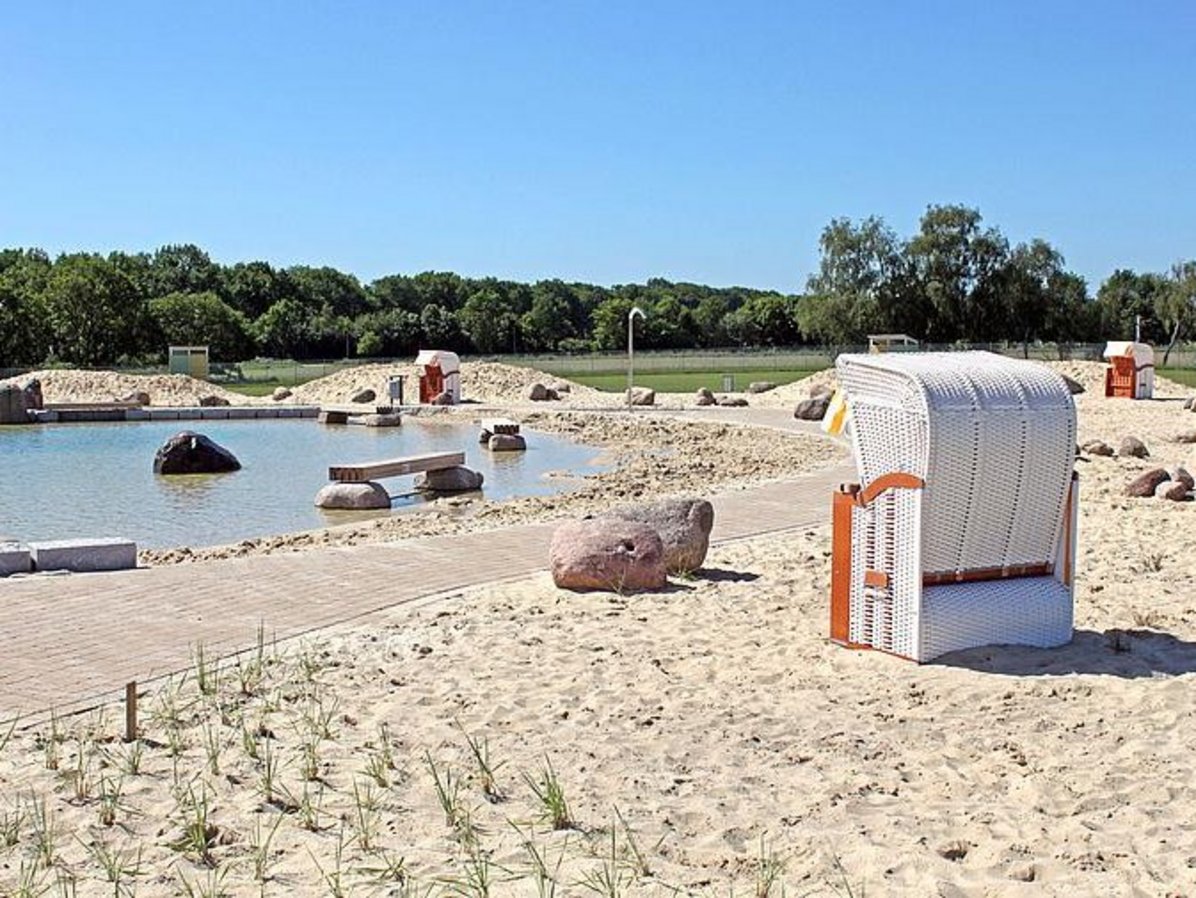 Hier ist ein Ausschnitt aus dem Naturbad Olfen zu sehen. Im Vordergrund ist der Sandstrand mit großen Steinen und einem Strandkorb zu erkennen. Im Hintergrund sieht man den Nichtschwimmerbereich des Bades mit einer Holzbank und Steinen. Abschließend sind im Hintergrund Wiese und viele Bäume zu sehen. Darüber strahlend blauer Himmel.