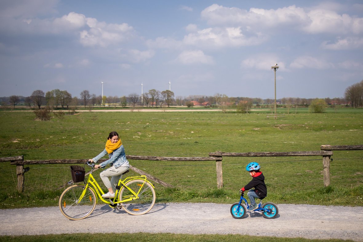 Eine Frau mit einem gelben Fahrrad und ein junge mit einem blauen Laufrad fahren auf dem Schotterweg in den Steverauen.