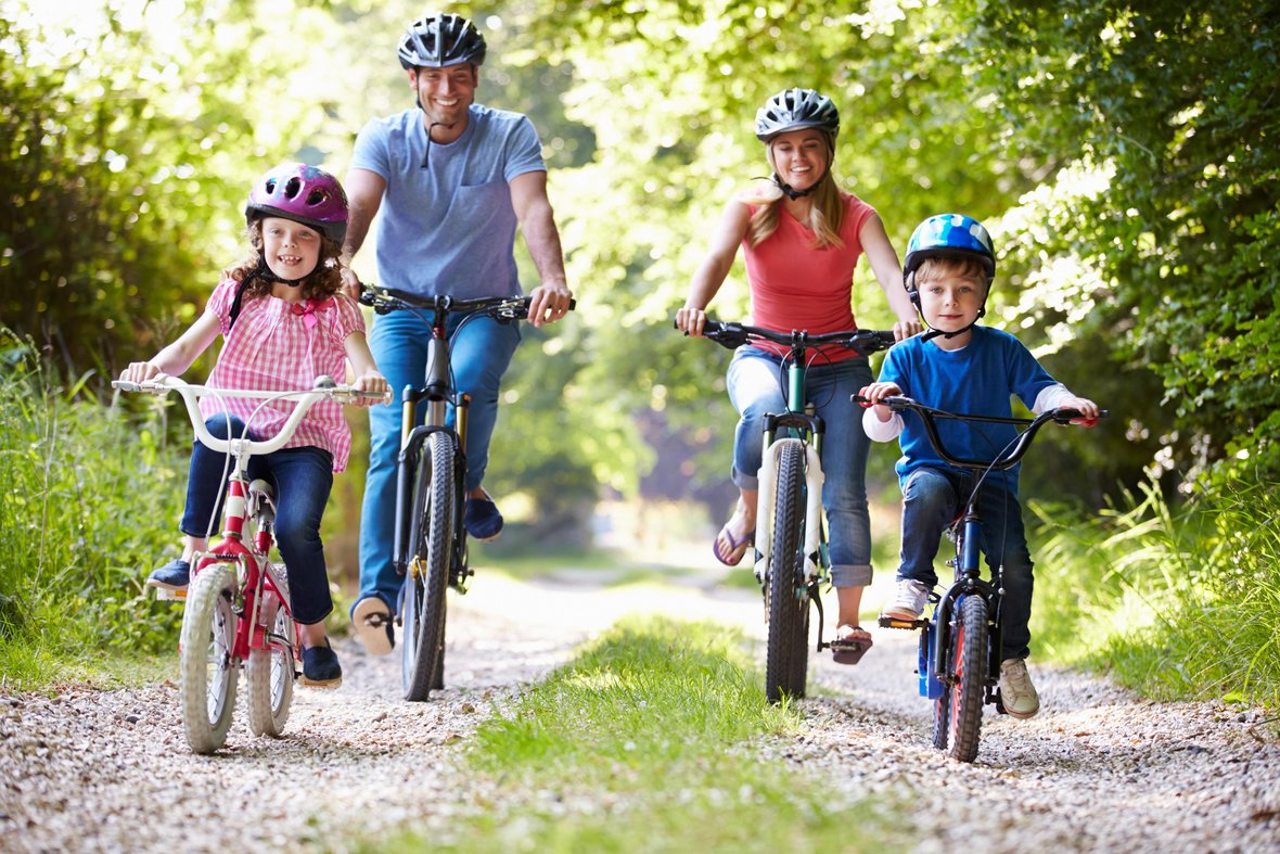 Eine Familie (Mutter, Vater, Sohn und Tochter) fährt über einen Schotterweg mit ihren Fahrrädern durch den Wald.