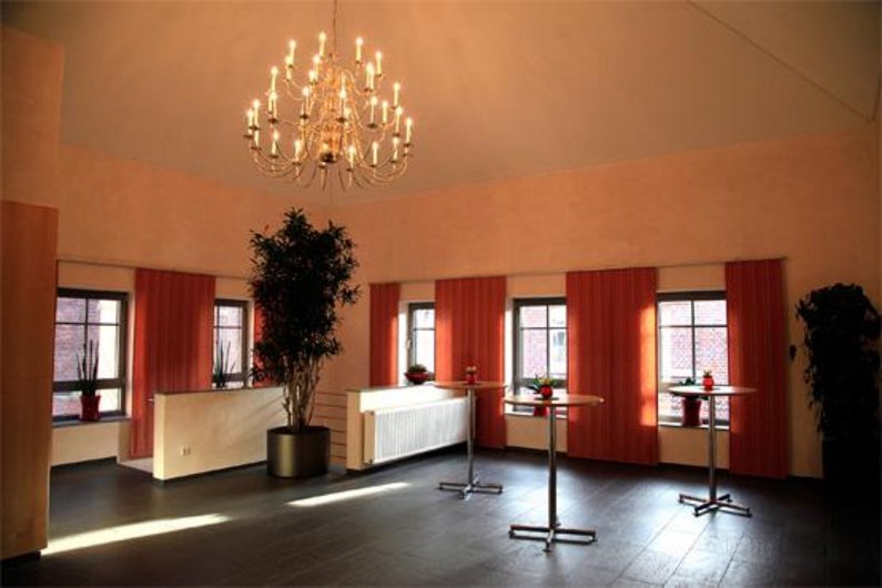 Im Foyer des Bürgerhauses hängen rote Gardinen, stehen drei Stehtische und großer Kornleuchter hängt von der Decke. Der Boden hat moderne schwarze Fliesen.