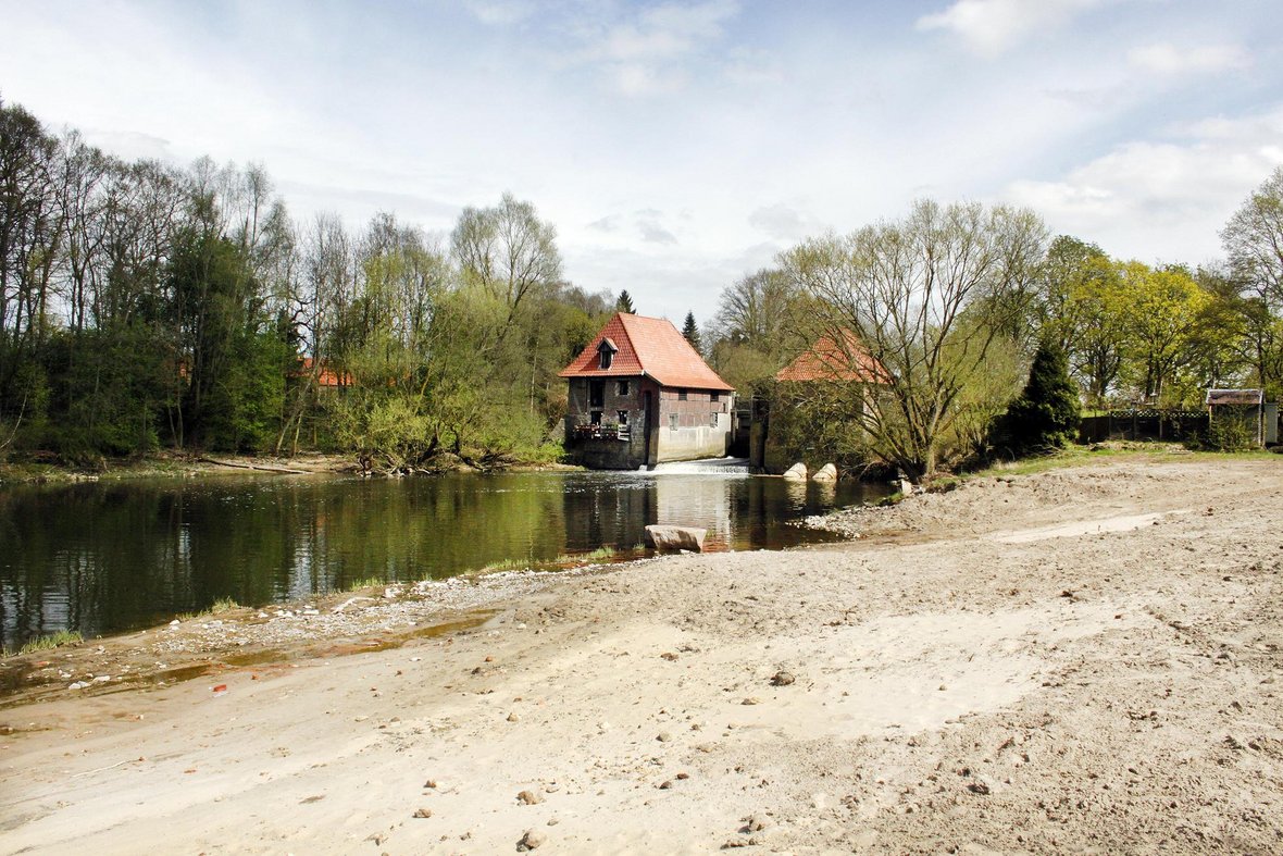 Im Vordergrund ist der Sandstrand des Flussstrandes zu sehen. Dahinter fließt die Stever. Im Hintergrund sind außerdem die beiden Gebäude der Füchtelner Mühle sowie viele grüne Bäume zu sehen.