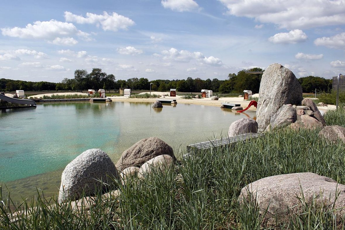 Auf dem Foto ist der Nichtschwimmerbereich des Naturbades zu sehen. Man erkennt im Vordergrund das grüne Schilf und im Hintergrund der Sandstrand mit Strandkörben zu erkennen.