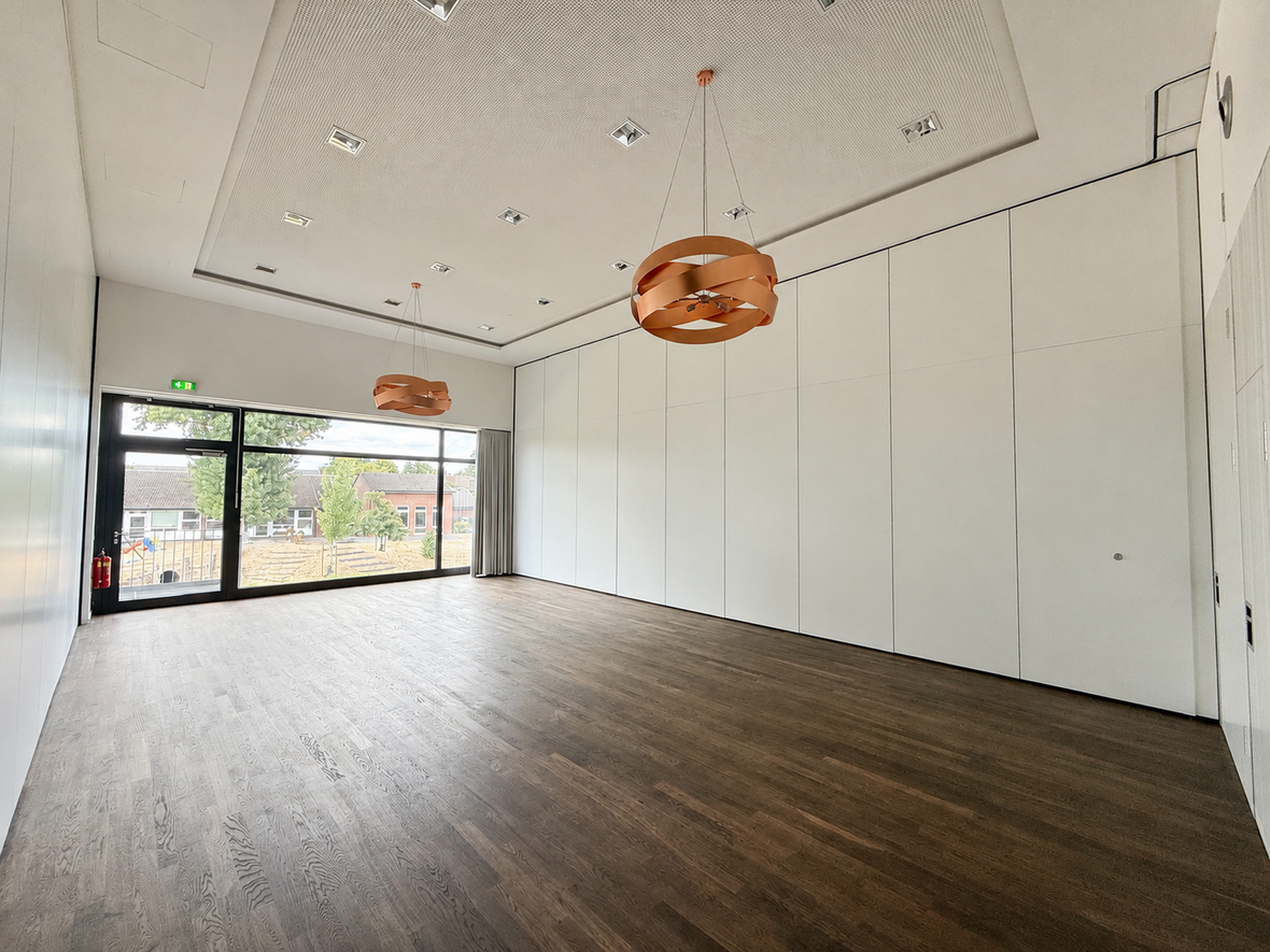 Heller, leerer Veranstaltungssaal mit dunklem Holzboden, weißen mobilen Trennwänden, großen Fenstern und zwei runden Pendelleuchten in Holzoptik.