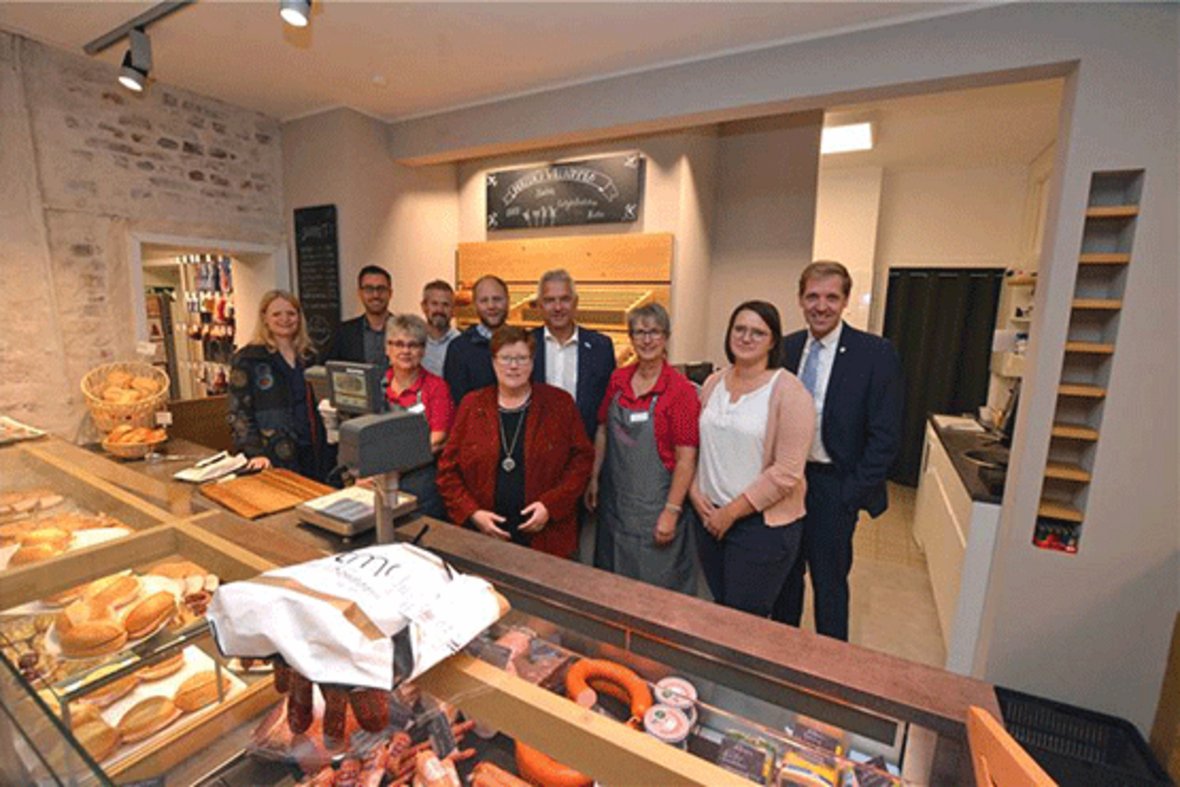 Bürgermeister Wilhelm Sendermann und Landrat Dr. Schulze Pellengahr stehen mit dem Team des Vinnumer Dorfladens hinter der Theke des Dorfladens. Im Vordergrund sind Back- und Wurstwaren zu sehen.