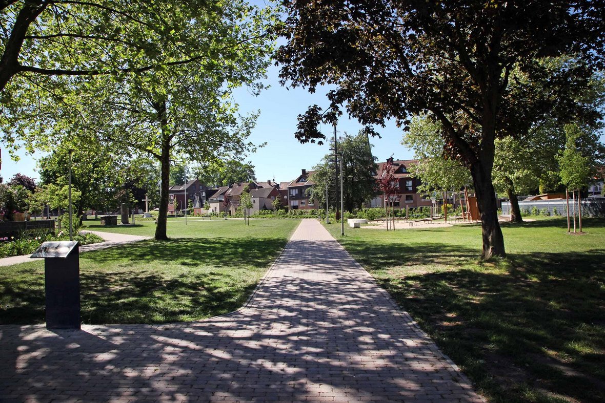 Zu sehen ist der gepflasterte Weg, der in den Olfener Stadtpark führt. Drumherum viele grüne Bäume und grüner Rasen. Außerdem ist im Hintergrund der Spielplatz zu erkennen.