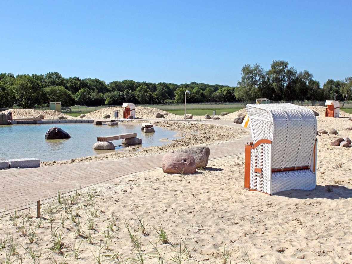 Auf dem Foto ist ein weißer Strandkorb im Sandstrand des Naturbades zu sehen. Im Hintergrund sieht man den Bereich des Nichtschwimmerbeckens.
