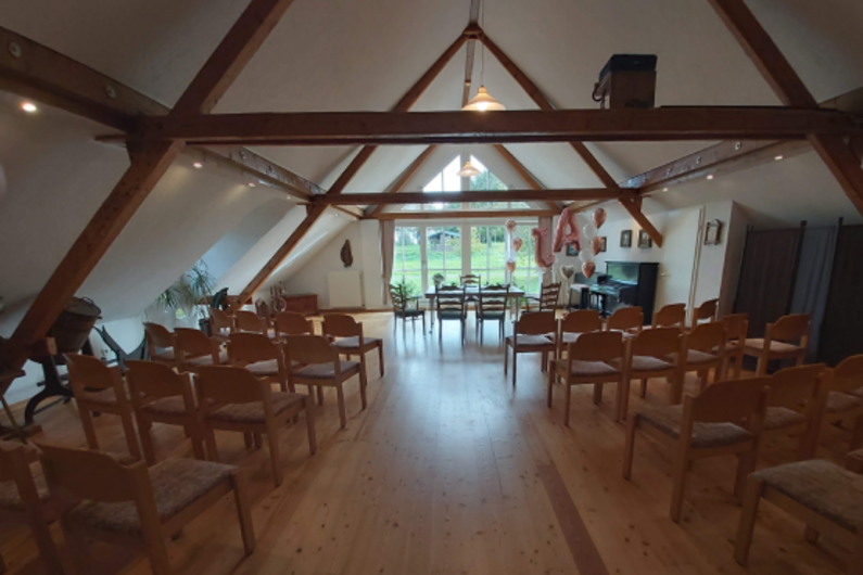 Das Trauzimmer des Heimathauses hat ein Spitzdach und alte Balken. Der Boden und die Stühle sind aus Holz, im gleichen Farbton wie die Balken. Durch die großen Fenster erscheint der Raum sehr hell.
