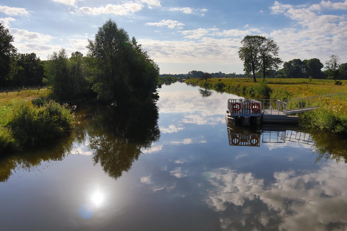 Mit Blick von der Kökelsumer Brücke schaut man auf den Floßanleger in der Steveraue. Das Floß ist unbesetzt, der Himmel spiegelt sich in der ruhigen Stever wieder und es ist ein sonniger Tag.