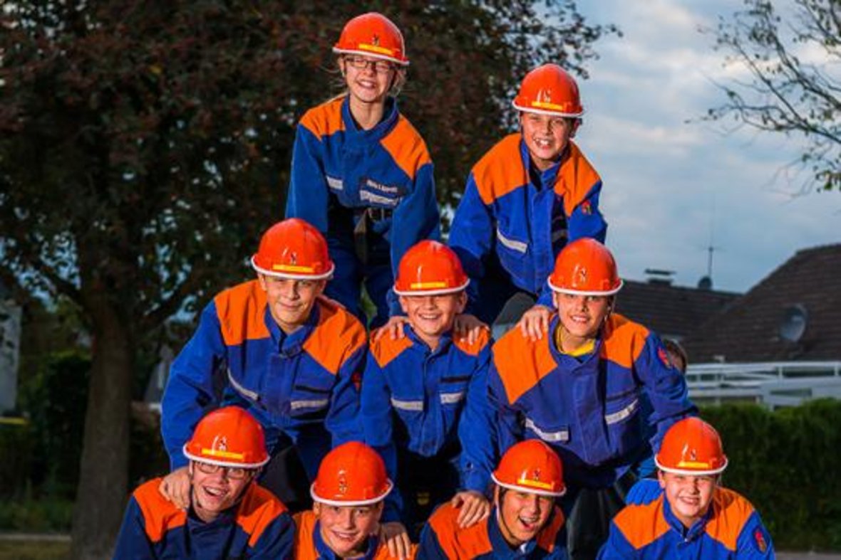 9 Kinder der Jugendfeuerwehr der freiwilligen Feuerwehr Olfen e. V. bilden auf diesem Foto eine Pyramide. 4 Kinder sind unten, 3 darüber und darüber nochmal 2 Kinder. Alle tragen blau-orangene Anzüge und orangene Helme. 