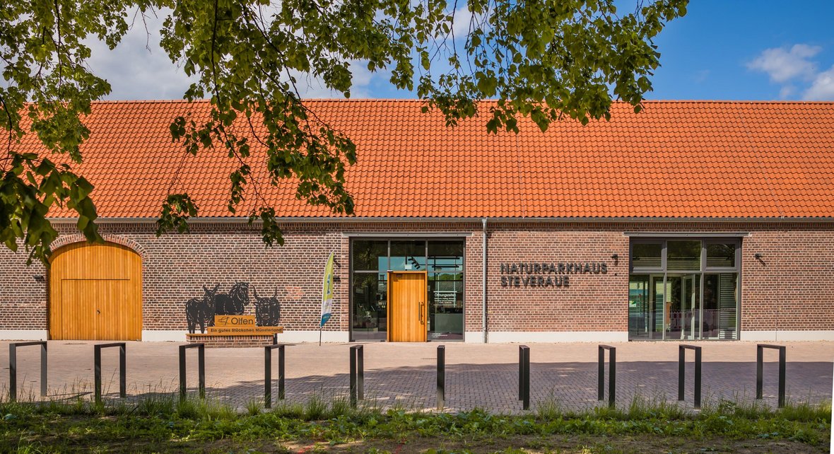 Das Foto zeigt die lange modernisierte Scheune in der sich das Naturparkhaus Steveraue befindet. Im Vordergrund sind schwarze Fahrradständer, links neben der Eingangstür ist der Fotopoint zu sehen und rechts daneben steht in großen Buchstaben NATURPARKHAUS STEVRAUE.