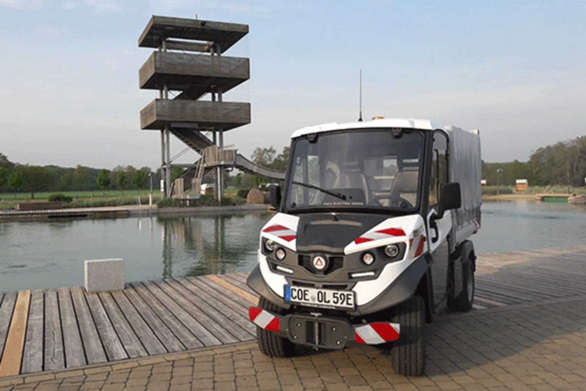 Ein kleines weißes E-Auto steht vor dem Schwimmerbecken des Naturbads Olfen.