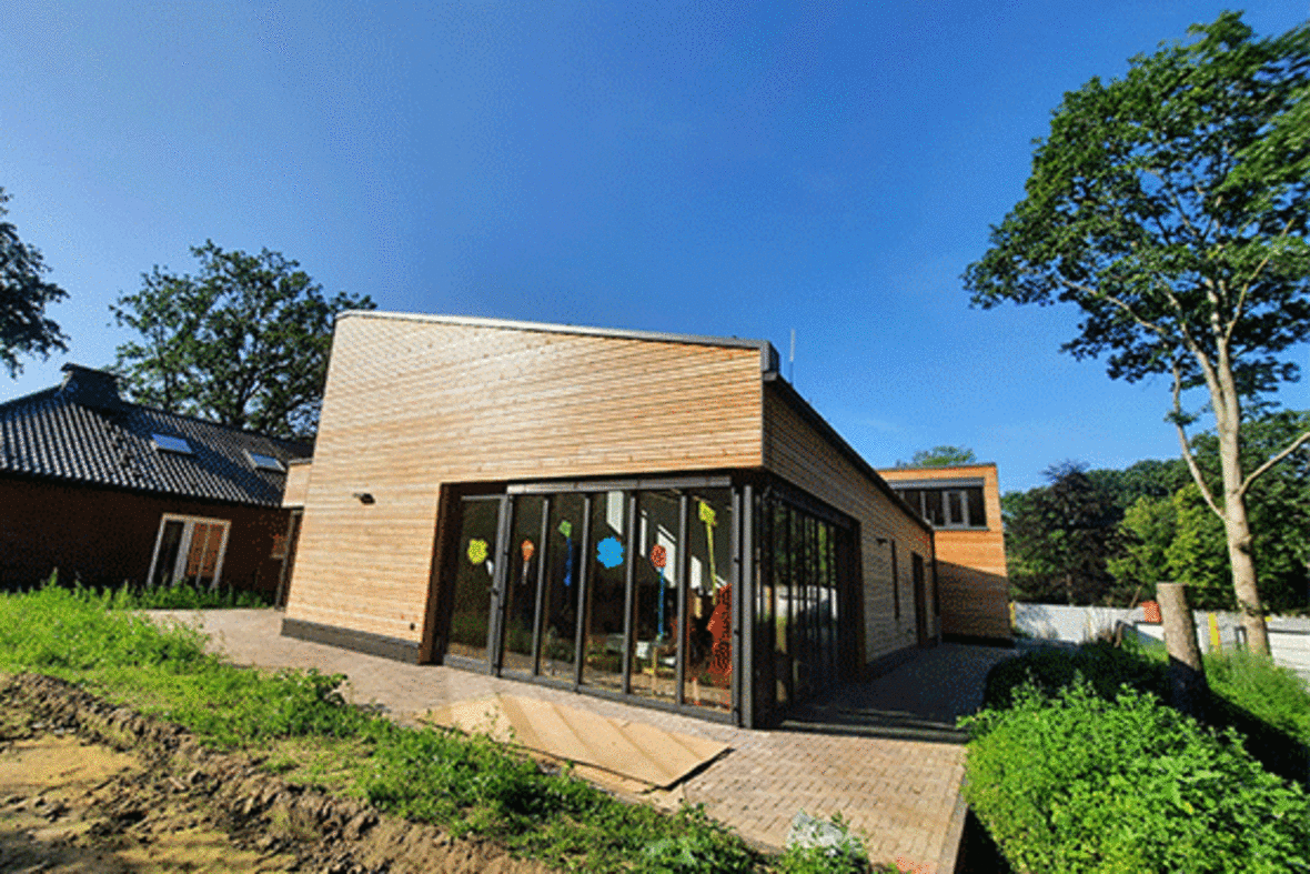 Hier sieht man den Naturkindergarten "An der Mühle". Der Bau ist modern aus Holz und hat große Scheibenfronten in den viele bunte gebastelte Bilder hängen.
