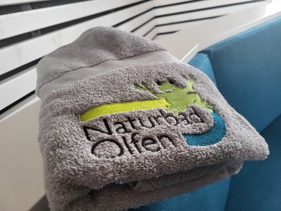 Hier ist das graue Handtuch mit dem bunten Logo "Naturbad Olfen" zu sehen.