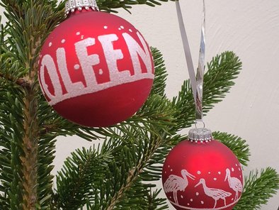 Zwei rote Weihnachtskugeln hängen am Weihnachtsaum. Mit weißem Glitzer ist dort die Aufschrift "Olfen" und das Motiv von Störchen zu sehen.