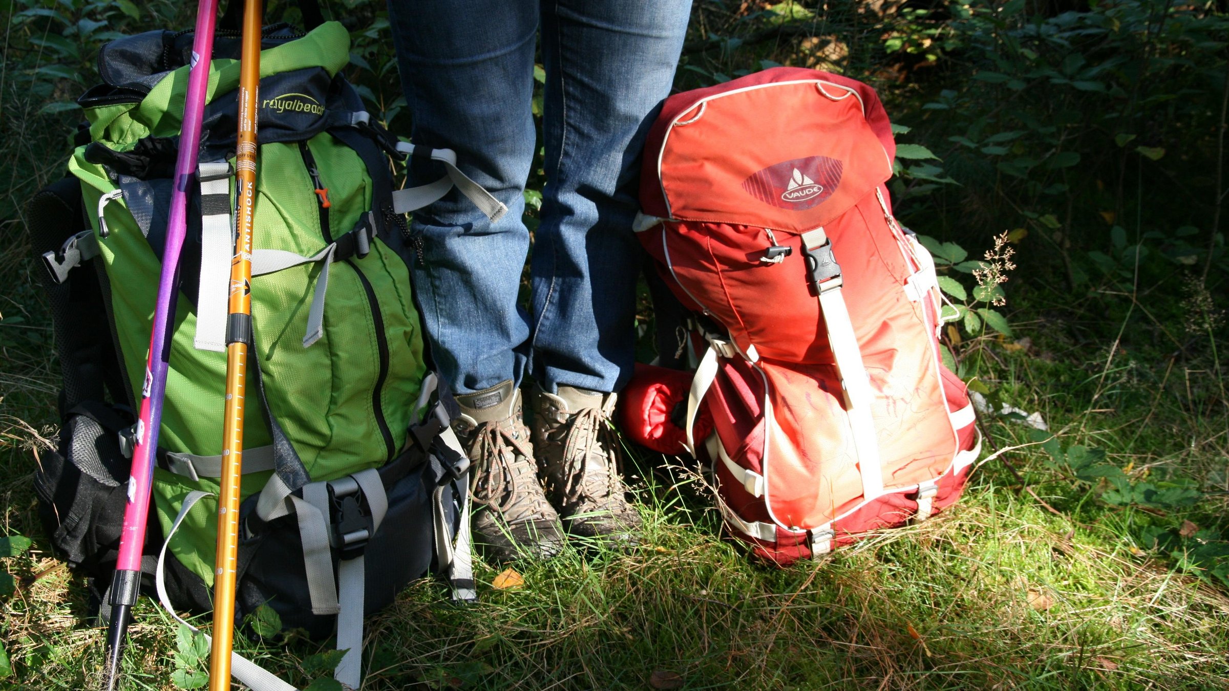 Beine einer Frau mit Wanderschuhen, daneben Wanderstöcke, und zwei Rücksäcke.