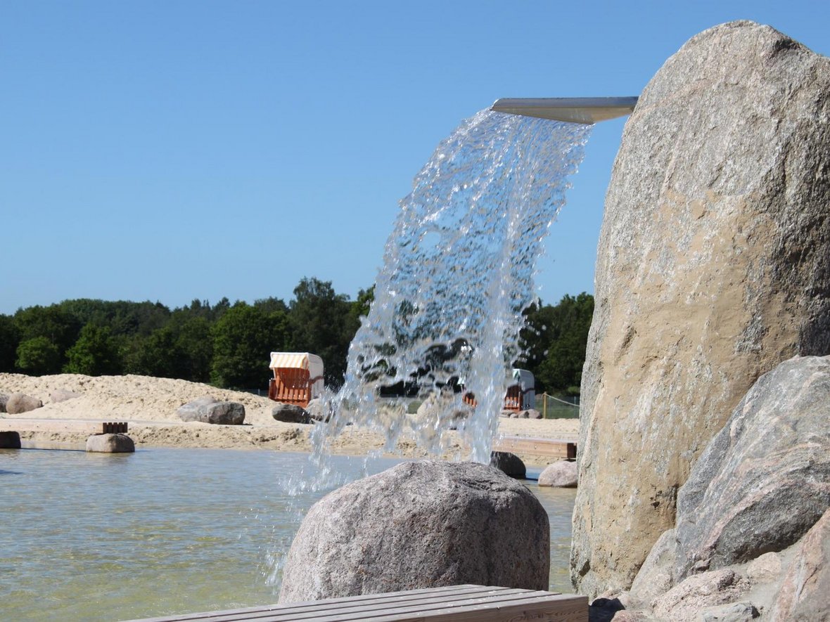 Der Ausschnitt des Naturbades in Olfen zeigt einen der großen Geysire aus dem Wasser strömt. Im Hintergrund sind der Nichtschwimmerbereich, der Sandstrand und ein Strandkorb zu erkennen.