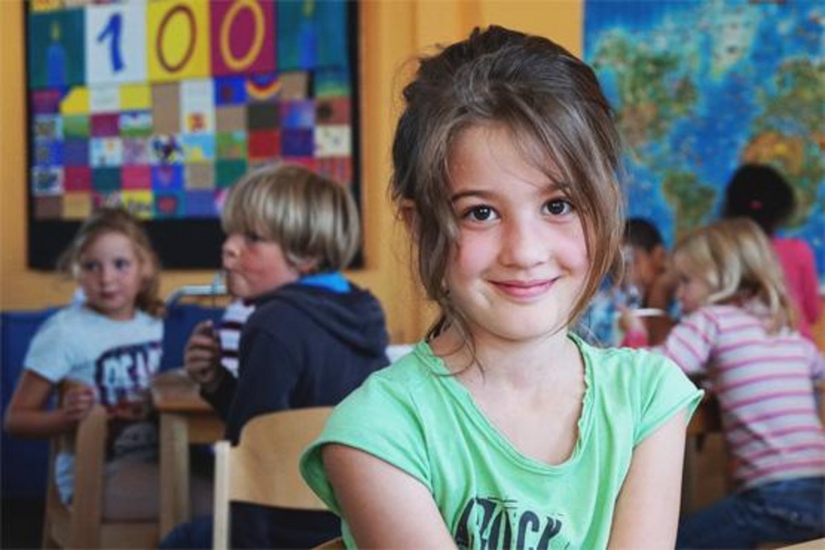 Auf dem Foto sind mehrere Kinder beim Essen an den Tischen in der OGS zu sehen. Im Vordergrund schaut ein Mädchen in die Kamera.
