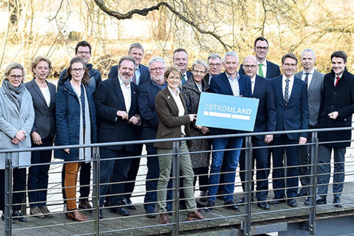 17 Personen aus dem Arbeitskreis 2Stromland stehen gemeinsam auf einer Brücke. Bürgermeister Wilhelm Sendermann hält ein Schild mit der Aufschrift 2Stromland hoch.