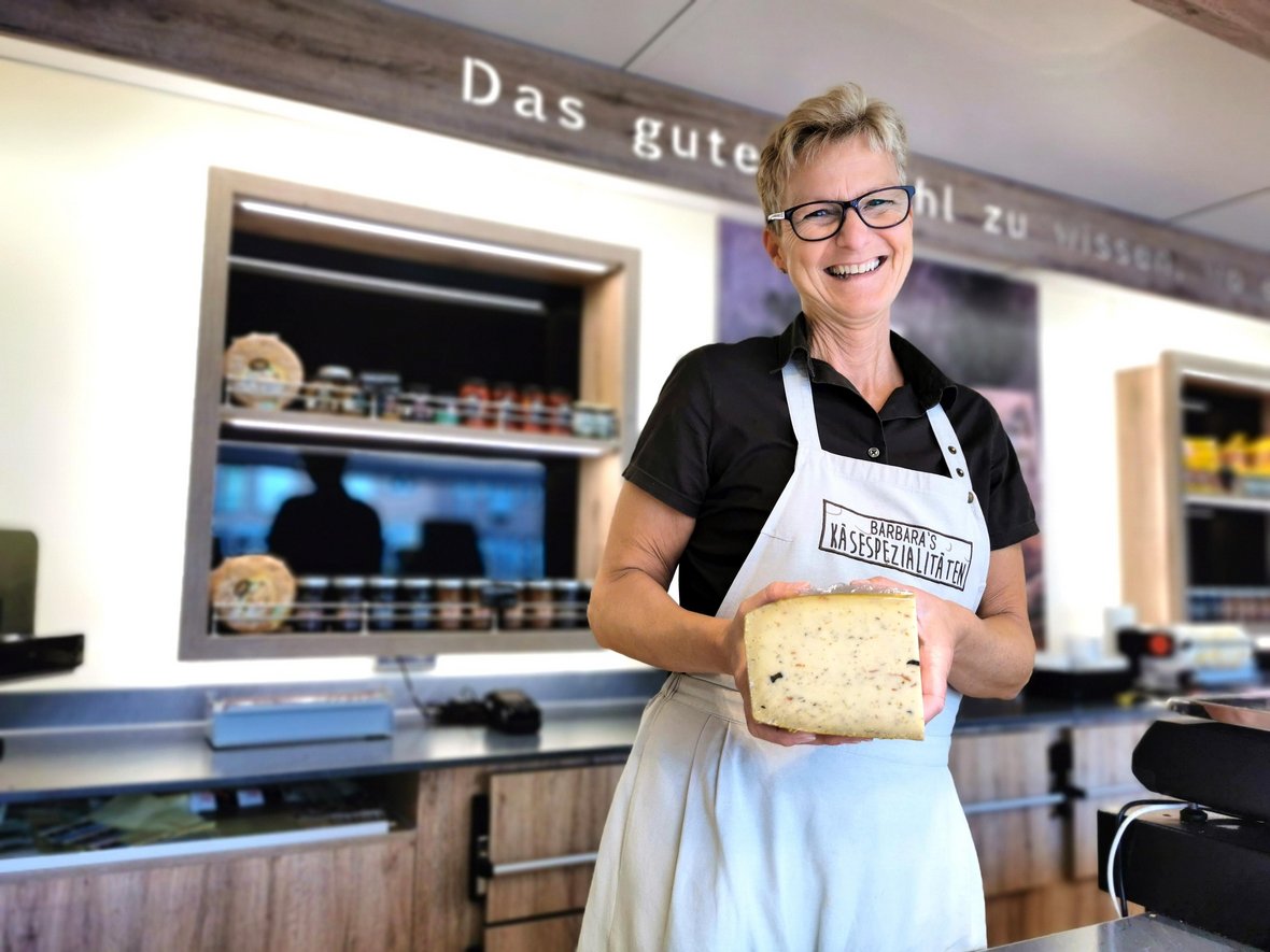 Barbara Otto vom Käsestand auf dem Wochenmarkt präsentiert ein Stück Käse in ihrem Verkaufswagen. Sie trägt eine Schürze mit der Aufschrift "Barbaras Käsespezialitäten".