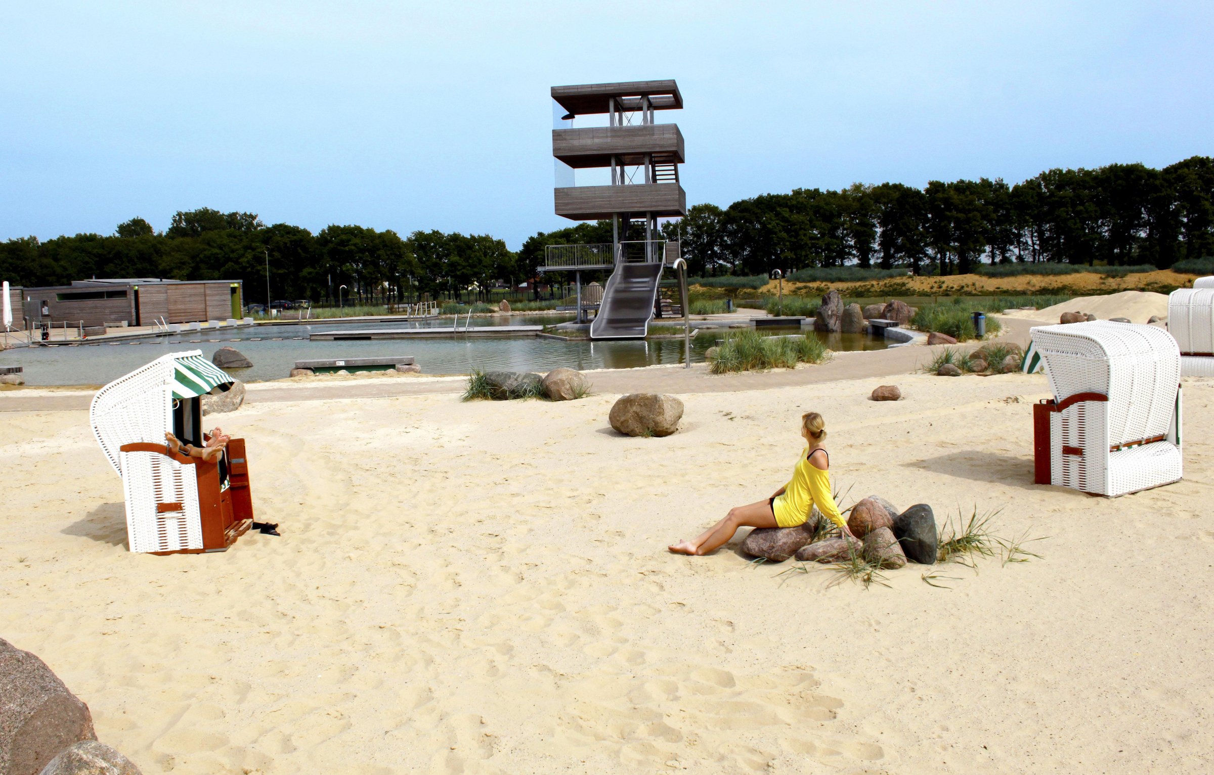Auf dem Foto sind der Strandbereich im Naturbad, der Spring- und Aussichtsturm und mehrere Strandkörbe zu sehen. Auf einem der Steine sitzt eine blonde Frau mit einem gelben Shirt.