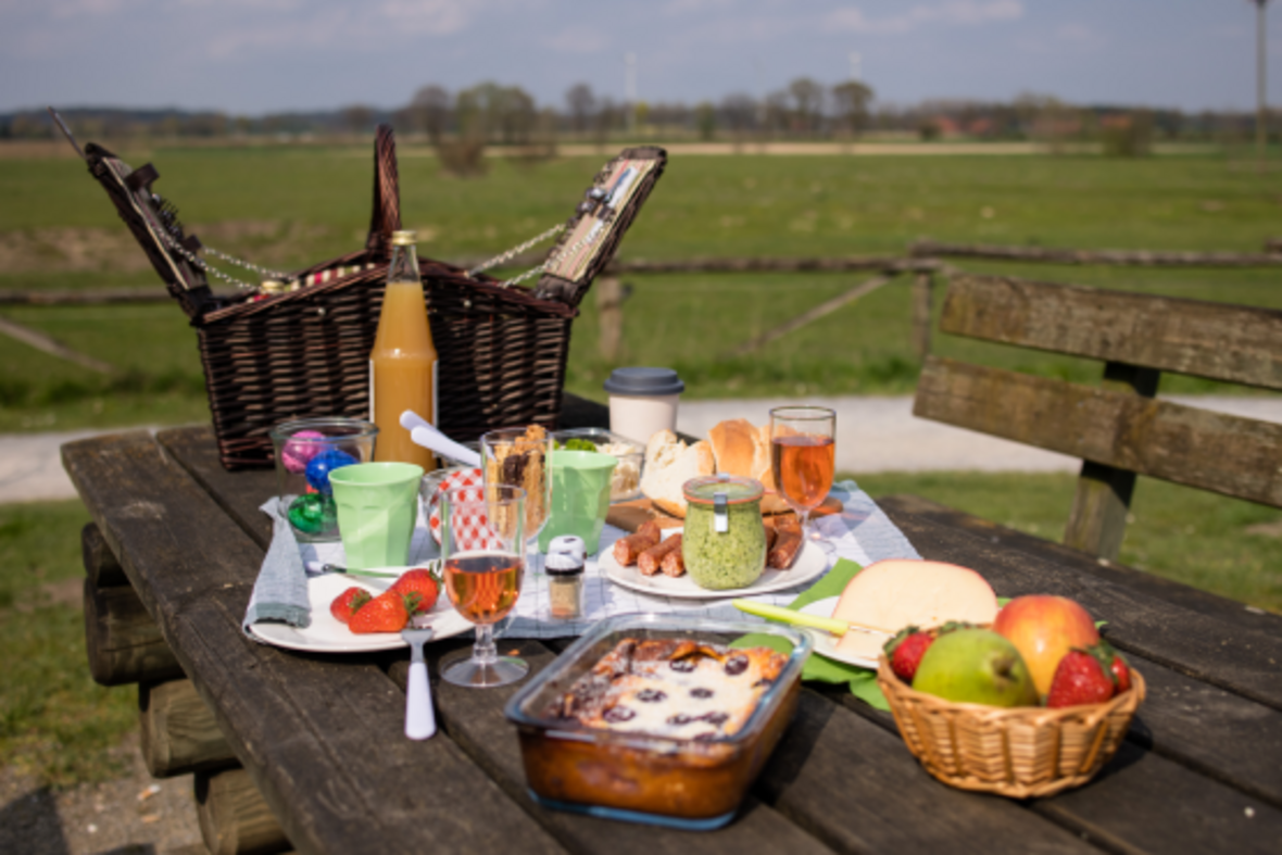 Auf einem Holztisch in den Steverauen steht ein großer geflochtener Picknickkorb. Davor sind viele Leckereien aus Obst, Gemüse und Brot angerichtet.