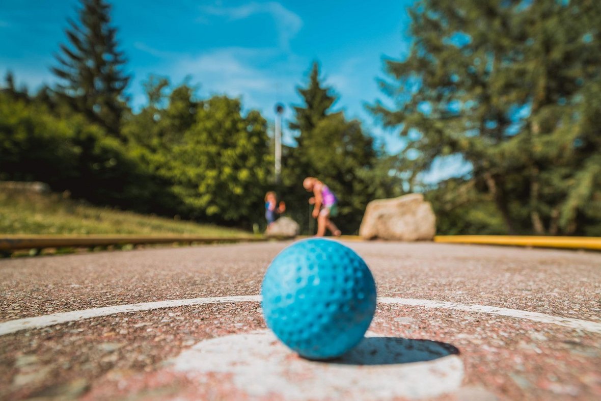 Ein hellblauer Minigolfball liegt im Vordergrund auf dem Startpunkt. Unscharf im Hintergrund erkennt man die beiden Spieler.
