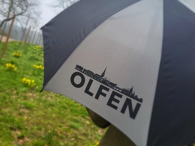 Der Regenschirm ist grau/schwarz. Auf einem der grauen Elemente steht in großer schwarzer Schrift "OLFEN", darüber ist eine "Skyline" von Olfen zu erkennen.