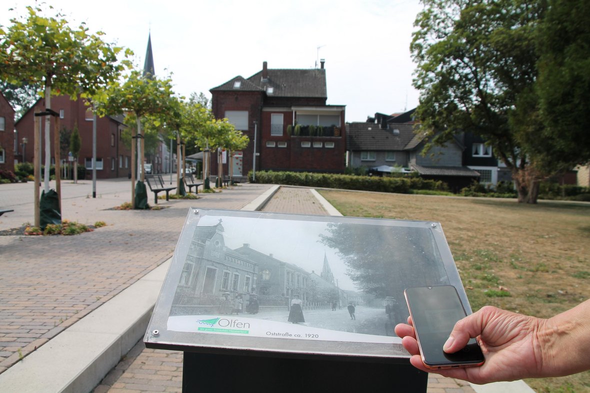 Hier ist eine schwarze Fotostele an der Oststraße zu sehen. Im Hintergrund sieht man den Stadtpark und die St. Vitus Kirche. Auf der Stele ist ein Foto der Oststraße aus demselben Blickwinkel vom Jahr 1920 zu sehen. Davor hält eine Person ihr Smartphone und scannt den dort befindlichen QR-Code um zum Digitalen Stadtrundgang des Heimatvereins in Olfen zu gelangen.