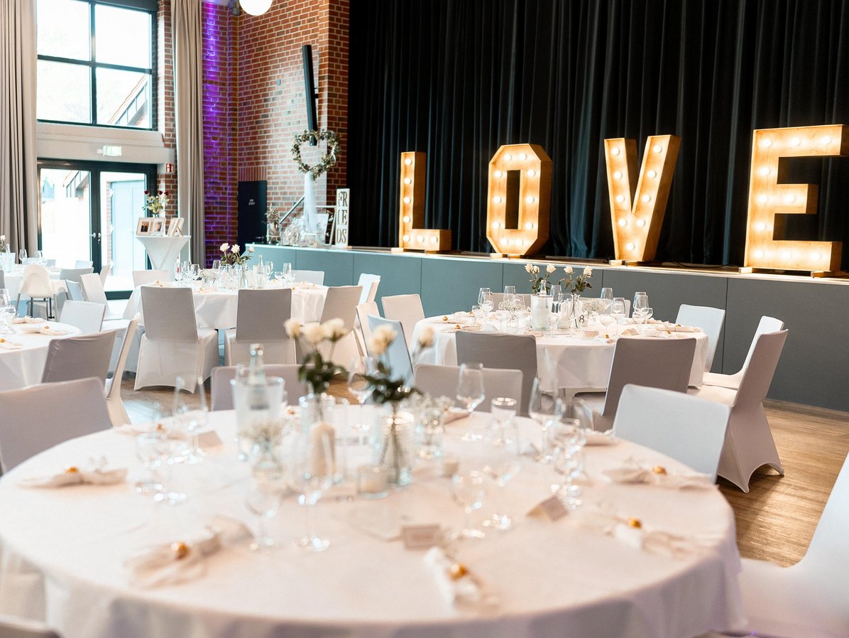 Festlich eingedeckter Saal der Stadthalle Olfen mit runden Tischen, weißen Stühlen und beleuchtetem „LOVE“-Schriftzug auf der Bühne.