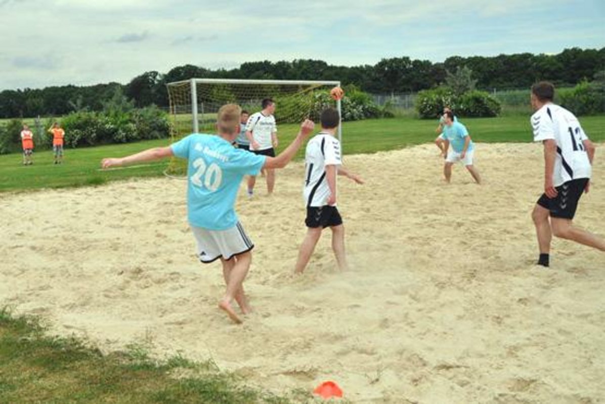 Auf einer größeren Sandfläche mit Fußballtoren sind mindestens 6 Spieler zu erkennen, die Fußball spielen.