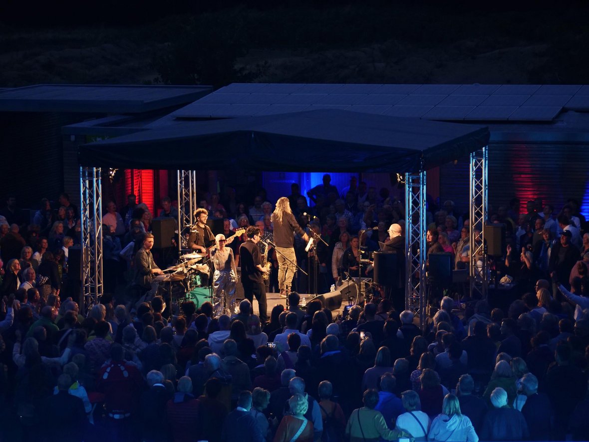 Im Fokus des Fotos steht die Bühne beim Open-Air-Konzert mit Wolf Coderas Session Possible im Naturbad Olfen. Drumherum stehen hunderte Menschen und schauen zur Bühne.