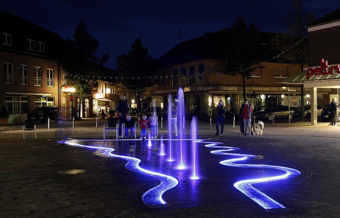 Der Brunnen am Marktplatz am Abend. Die Fontänen und Wasserrillen leuchten in einem hellen blau, drumherum stehen einige Personen und schauen zum Brunnen.