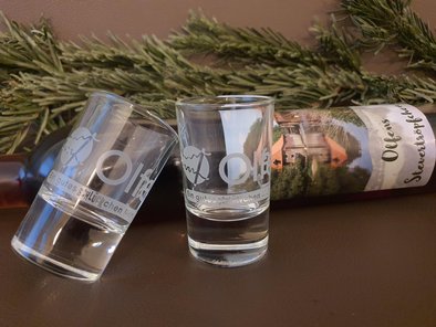 Auf dem Foto sind zwei Pinnchen/Schnapsgläser mit der Aufschrift "Ein gutes Schlückchen Münsterland" zu sehen. Im Hintergrund liegt eine Flasche "Olfens Stevertröpfchen".