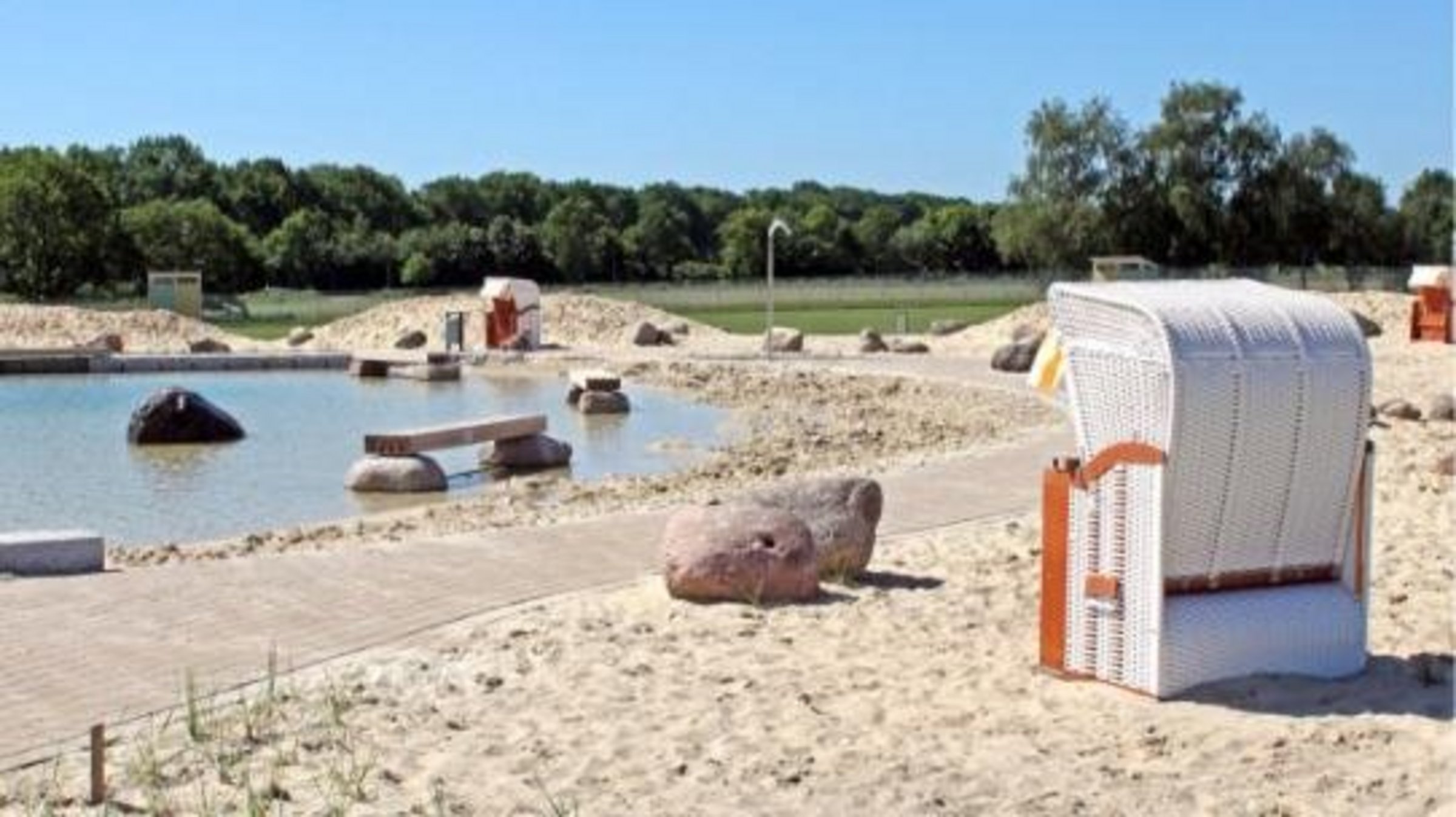 Auf dem Sandstrand vom Naturbad Olfen steht ein großer weißer Strandkorb. Im Hintergrund ist der Nichtschwimmerbereich mit Holzbänken und Steinen zu sehen.