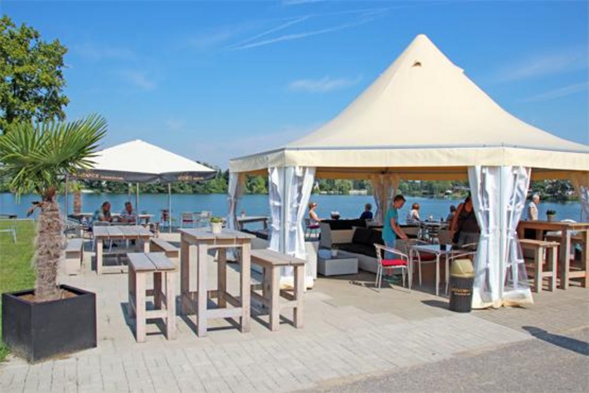 Hier wird der Gastronomie-Bereich vom Ternscher See gezeigt. Im Hintergrund sieht man das Wasser des Ternscher Sees. Auf der gepflasterten Fläche im Vordergrund sind weiße große Pagoden zu sehen unter denen Tische zum Essen und Verweilen aufgestellt wurden.