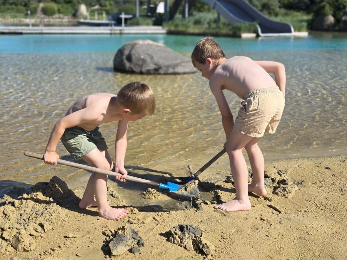 Zwei Kinder buddeln im Sand des Nicht-Schwimmer-Bereichs im Naturbad Olfen. Im Hintergrund ist der Sprung- und Aussichtsturm des Bades zu sehen.