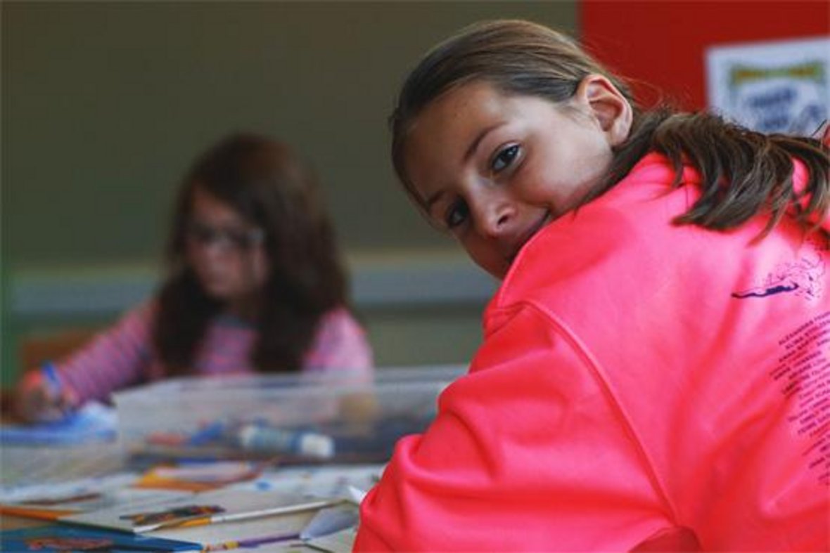 Zwei Kinder sitzen am Tisch und machen Hausaufgaben. Das Mädchen im Vordergrund schaut über die Schulte in die Kamera.
