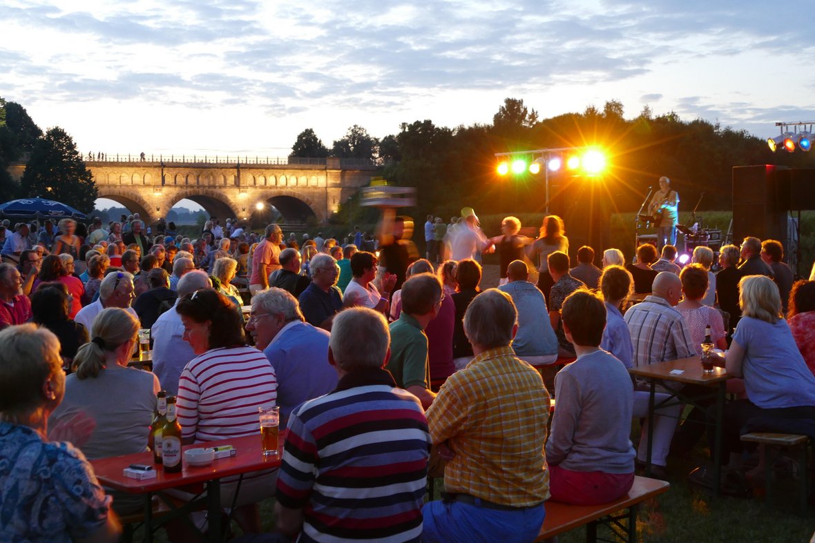 Das Foto zeigt hunderte Menschen auf einem der Summer Special Abende am Steverstrand in Olfen. Auf der Bühne sieh man einen Sänger mit seiner Gitarre und im Hintergrund die beleuchtete Dreibogenbrücke.