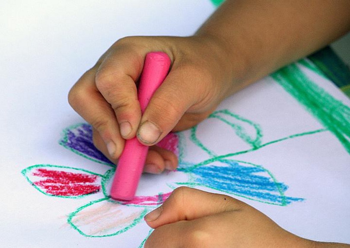 Auf dem Foto sind Kinderhände zu sehen, die mit einem pinken Wachsmalstift auf einem Blatt Papier zeichnen.