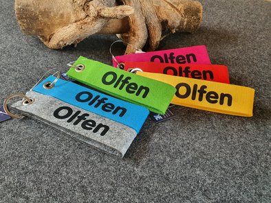 Schlüsselanhänger in verschiedenen Farben (grau, blau, grün, gelb, rot, pink) aus Filz mit der Aufschrift "Olfen" in schwarz.