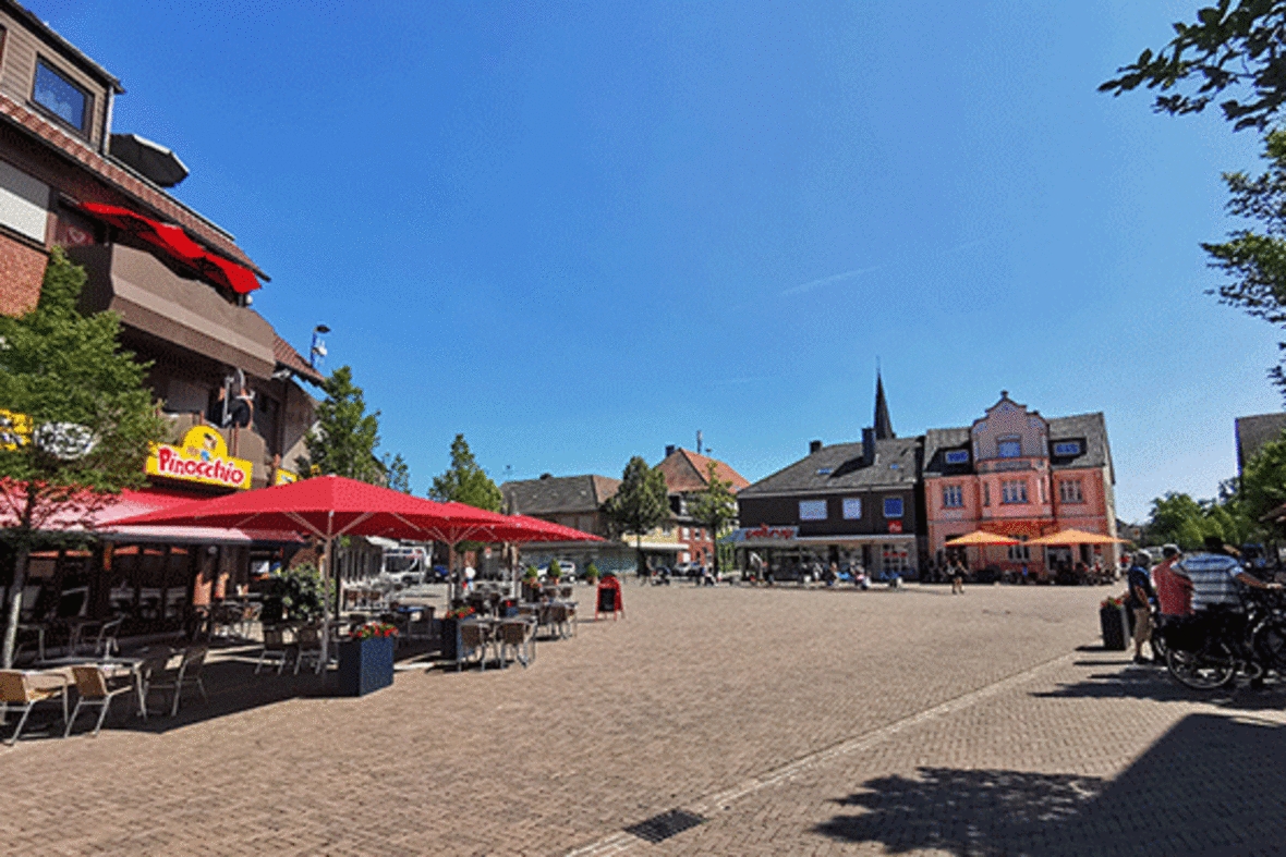 Hier ist der Marktplatz aus Sicht der Funnenkampstraße zu sehen.  Bei der Pizzeria Pinocchio sind rote Sonnenschirme aufgespannt. Die Kirchturmspitze ist noch zu erkennen. Der Himmel ist strahlend blau.